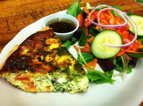 A slice of vegetable quiche served with a fresh salad of greens, tomatoes, cucumbers, and a small cup of dressing.