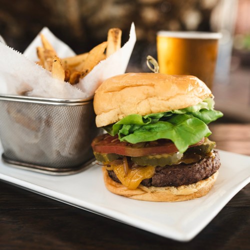 A juicy burger with lettuce, tomato, pickles, and cheese, served with a side of fries and a glass of beer.