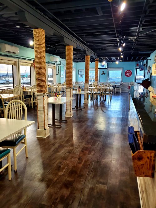 Interior of a restaurant featuring wooden floors, tables, and rope-wrapped columns, with large windows and a bar area.