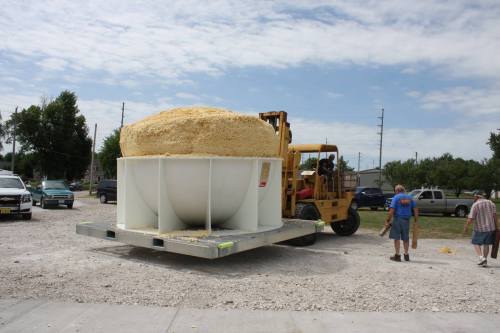 A large white container filled with yellow material is being loaded onto a flatbed truck by a forklift.