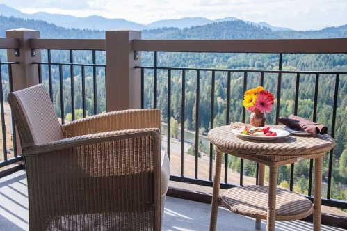 A cozy balcony with a chair and a table, featuring a plate of fruit and flowers, overlooking a scenic mountain view.