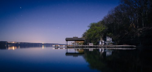 A serene lakeside scene at night, featuring a dock and trees reflecting on calm water under a starry sky.