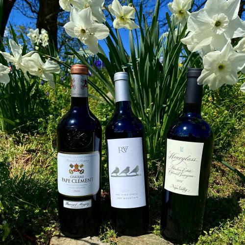 Three wine bottles displayed in a garden with blooming white flowers and a clear blue sky in the background.