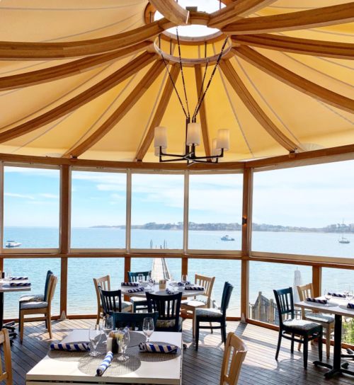 Bright dining area with wooden chairs and tables, featuring large windows overlooking a serene waterfront view.