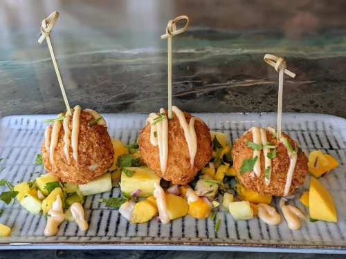 Three crispy fried balls drizzled with sauce, served on a bed of diced mango, pineapple, and herbs on a rectangular plate.