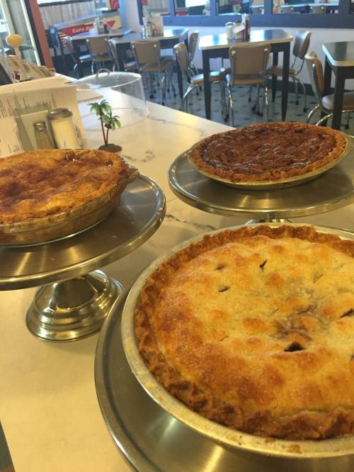 Three delicious pies on display: a golden apple pie, a chocolate pie, and a pecan pie, all on silver stands.