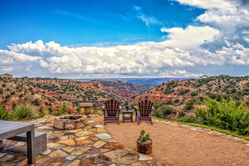 A scenic view of a canyon from a patio with two chairs, surrounded by greenery and dramatic clouds in the sky.