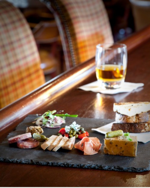 A slate platter with assorted meats, cheeses, pickles, and garnishes, alongside a glass of whiskey on a wooden table.