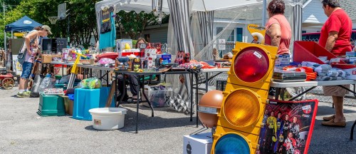 A bustling outdoor market with various tables displaying toys, tools, and colorful items under tents.