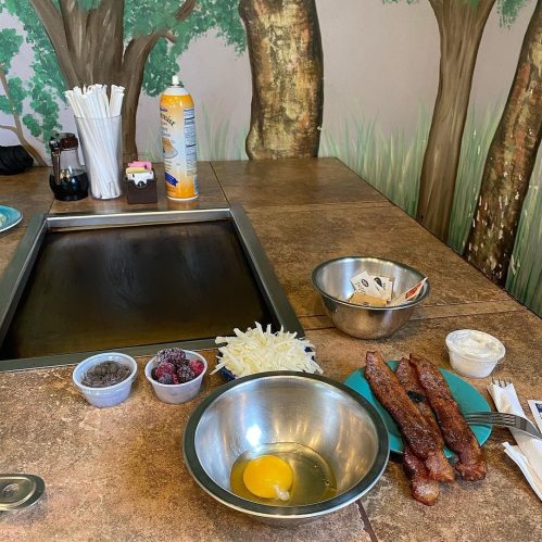 A table with a grill, bowls of ingredients including an egg, bacon, and berries, set in a nature-themed restaurant.