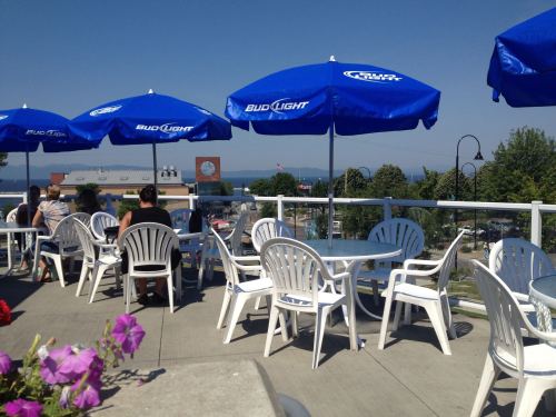 Outdoor dining area with white chairs and blue umbrellas, overlooking a scenic view of the water and distant trees.
