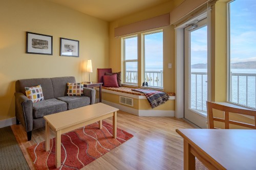 Cozy living area with a gray sofa, wooden table, and large windows overlooking a scenic view of water.