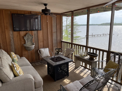 Cozy screened porch with a sofa, chairs, and a fire pit, overlooking a lake with a dock in the background.