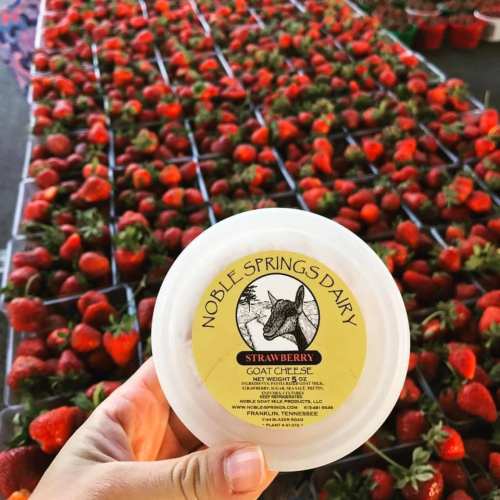 A hand holding a container of strawberry goat cheese in front of a display of fresh strawberries.