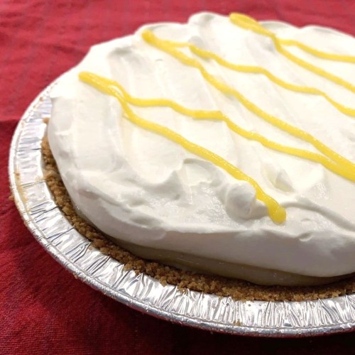 A creamy pie with a graham cracker crust, topped with whipped cream and drizzled with yellow sauce, on a red background.