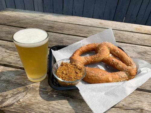 A large pretzel with sesame seeds served with mustard, alongside a glass of light beer on a wooden table.