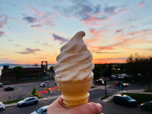A hand holds a soft-serve ice cream cone against a colorful sunset sky.