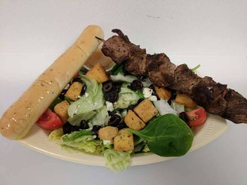 A plate with a fresh salad topped with olives, croutons, and a skewer of grilled meat, alongside a breadstick.
