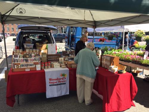 A vendor at a market selling various products, with a table covered in items and a car parked nearby.