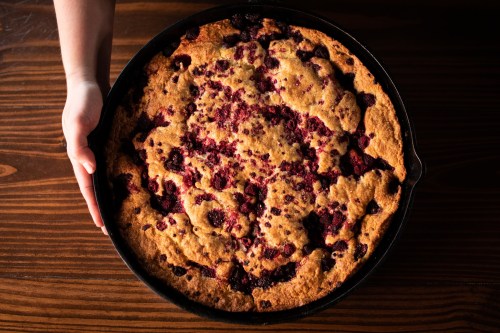 A hand holds a large, baked dessert with a golden-brown top and scattered berries, resting in a cast iron skillet.
