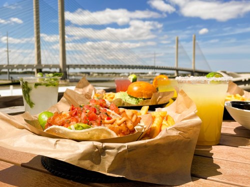 A vibrant outdoor dining scene featuring tacos, a burger, fries, and colorful drinks with a bridge in the background.