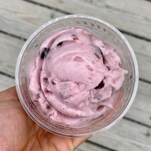 A hand holding a cup of pink berry sorbet, with a smooth texture and small fruit pieces, against a wooden background.