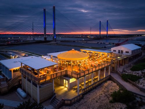 A waterfront restaurant illuminated at dusk, with a bridge and colorful sunset in the background.
