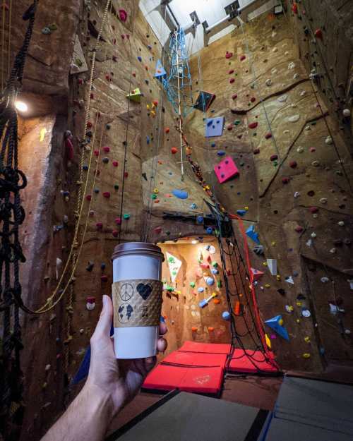 A hand holds a coffee cup in front of a colorful indoor rock climbing wall with climbing holds and ropes.