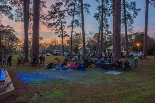 A crowd gathers outdoors under tall trees at sunset, enjoying an event with blankets and chairs set up on the grass.