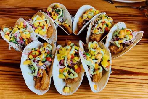 A platter of tacos filled with fried fish, colorful slaw, and drizzled with a creamy sauce, arranged on a wooden table.