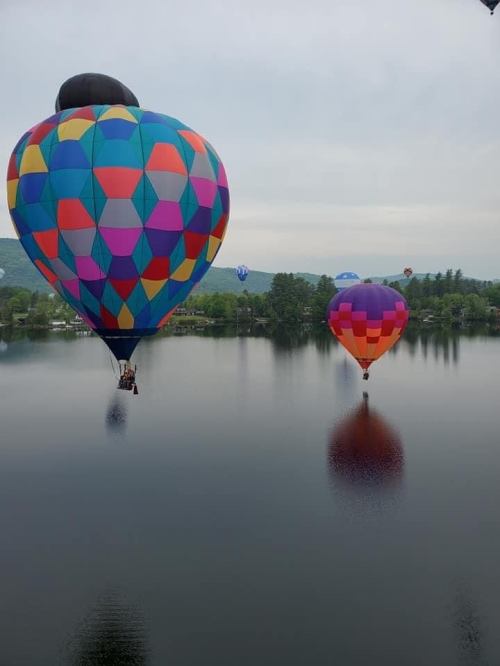 Two colorful hot air balloons float above a calm lake, reflecting on the water's surface under a cloudy sky.
