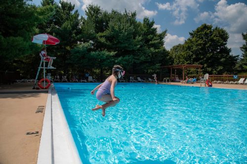 A child in a swimsuit jumps into a bright blue swimming pool on a sunny day, surrounded by trees and lounge chairs.