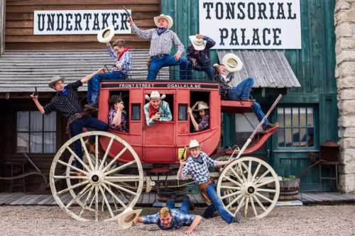 A group of cowboys in plaid shirts and hats poses playfully on a vintage stagecoach in a western-themed setting.