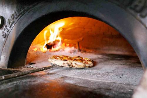 A pizza baking in a wood-fired oven, with flames and glowing embers visible in the background.