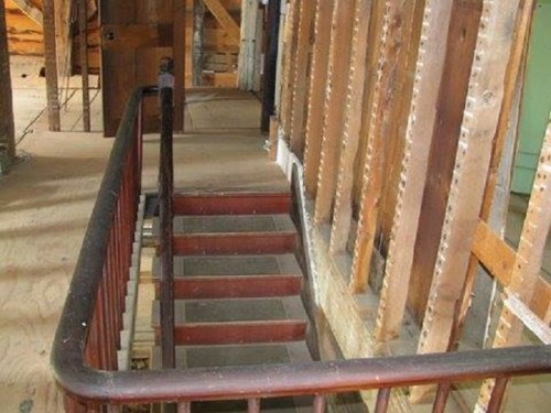 A wooden staircase with a dark railing, leading down in a partially constructed interior space with exposed walls.