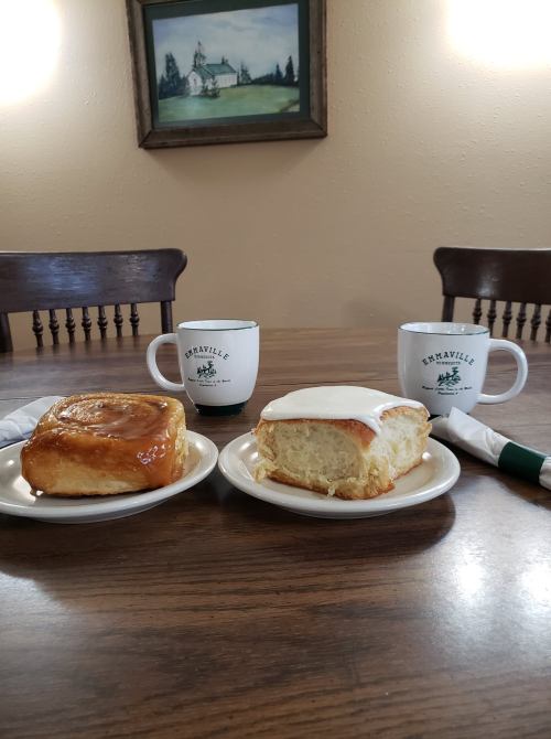 Two plates with cinnamon rolls, one glazed and one with icing, on a wooden table with coffee mugs in the background.