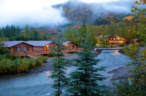 A serene landscape featuring wooden cabins by a river, surrounded by trees and misty mountains at dusk.