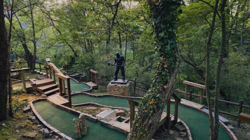 A mini-golf course surrounded by trees, featuring a statue and winding paths on a green carpet.