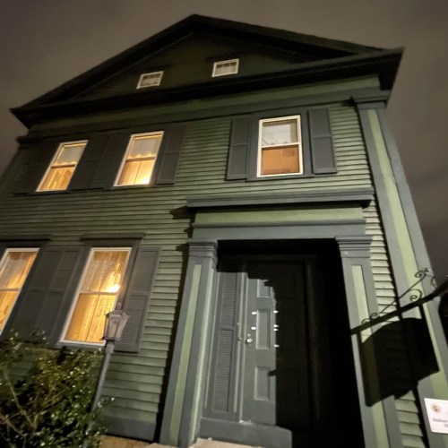 A tall, dark green house at night, with illuminated windows and a welcoming front door.
