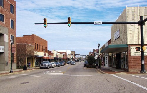A quiet downtown street with shops, parked cars, and a traffic light showing green under a cloudy sky.