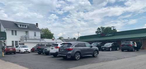 A parking lot with several cars, a two-story house, and a green-roofed building under a cloudy sky.