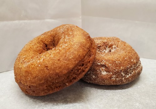 Two freshly baked donuts, one coated in sugar and the other plain, resting on a white paper background.