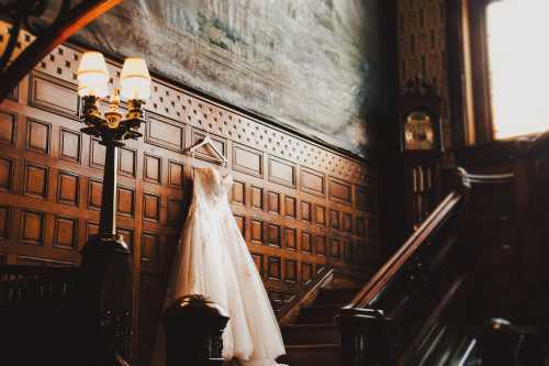 A wedding dress hangs elegantly on a wooden wall near a staircase, illuminated by soft lamp light.