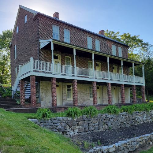 Historic brick building with a large porch, surrounded by greenery and stone landscaping.