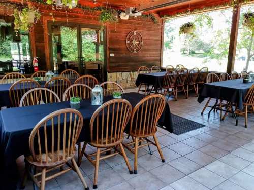 A spacious dining area with wooden chairs and black tablecloths, surrounded by greenery and large windows.