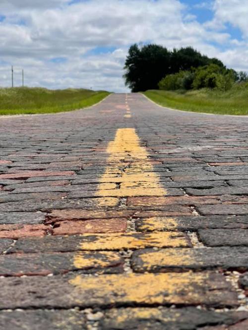 A close-up view of a brick road with a yellow centerline, leading towards a distant horizon under a cloudy sky.