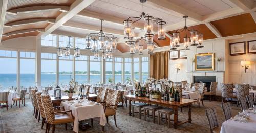 Elegant restaurant interior with large windows, ocean view, and tables set for dining. Warm lighting and stylish decor.