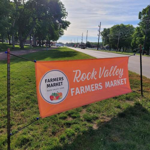 Banner for Rock Valley Farmers Market, featuring bright orange background and logo, set against a grassy area and road.