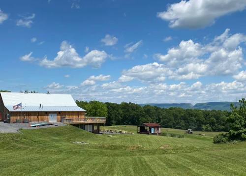 A scenic view of a wooden building with a deck, surrounded by green fields and rolling hills under a blue sky with clouds.