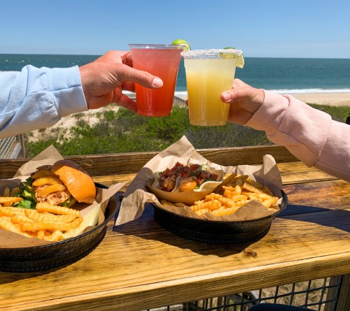 Two hands clink colorful drinks over a table with burgers and fries, with a beach and ocean in the background.